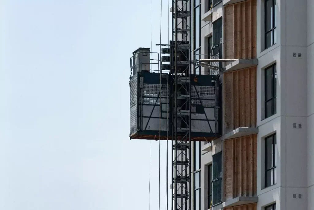 Építési felvonó egy modern épület homlokzatára szerelve, amely a teherlift bérlésének fontosságát mutatja be az építőiparban.