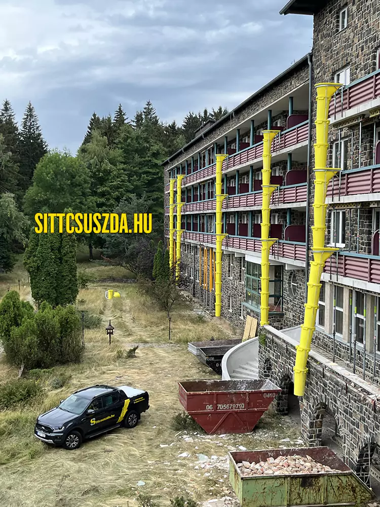 Sárga sitt csúszda rendszer egy többszintes épület homlokzatán felújítás közben, a törmeléket a lenti konténerekbe juttatva, bemutatva a törmelékledobó bérlését.